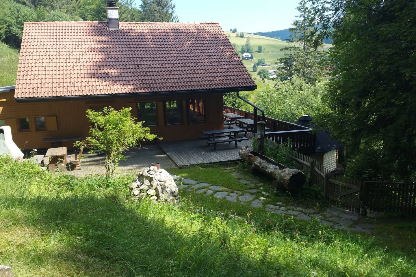 Terrasse des Berghauses mit Bänken und Holzbrunnen, im Hintergrund das Tal